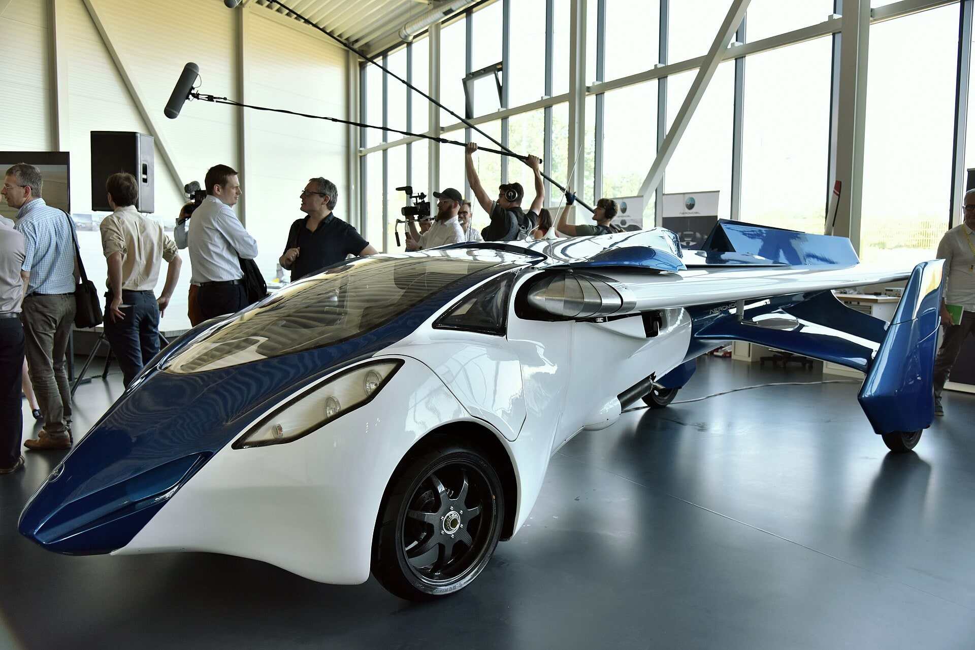 A white and blue AeroMobil flying car with its wings spread, in a glass-walled hall, watched by a camera crew.