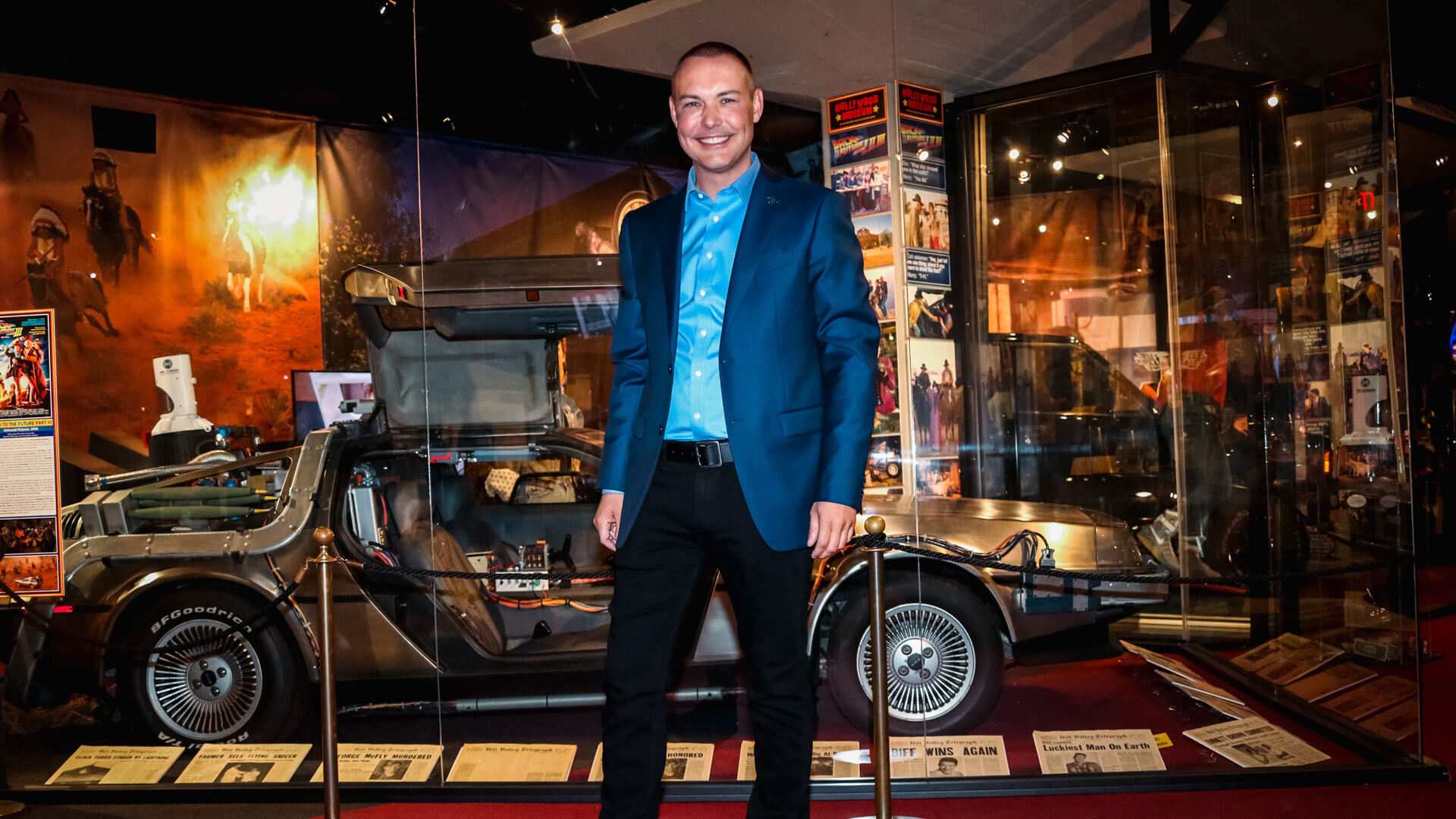 A man in a blue suit stands beside a DeLorean car in a museum display of film memorabilia.