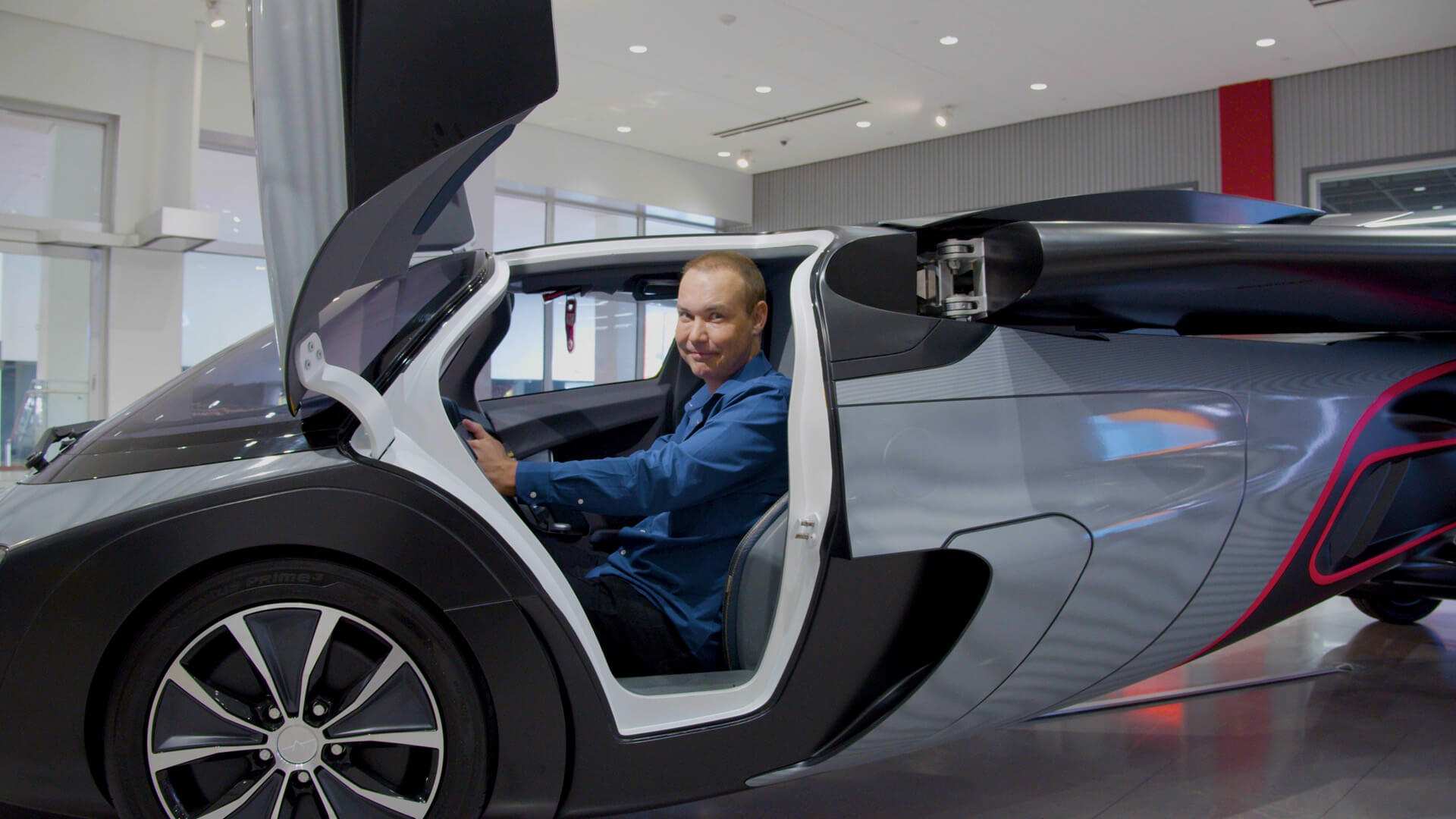 A man sits at the wheel of a silver vehicle with its door raised, in a showroom.