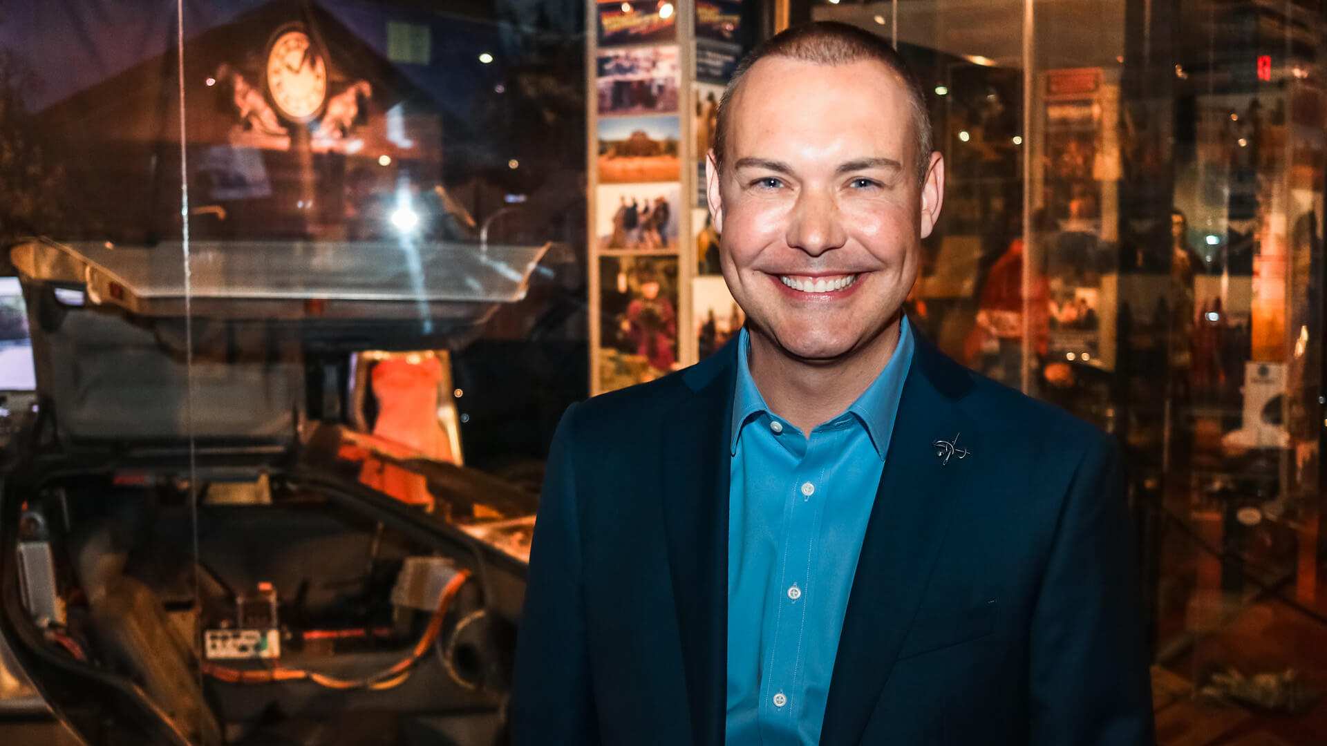 A man in a dark suit smiles in front of museum display cases.