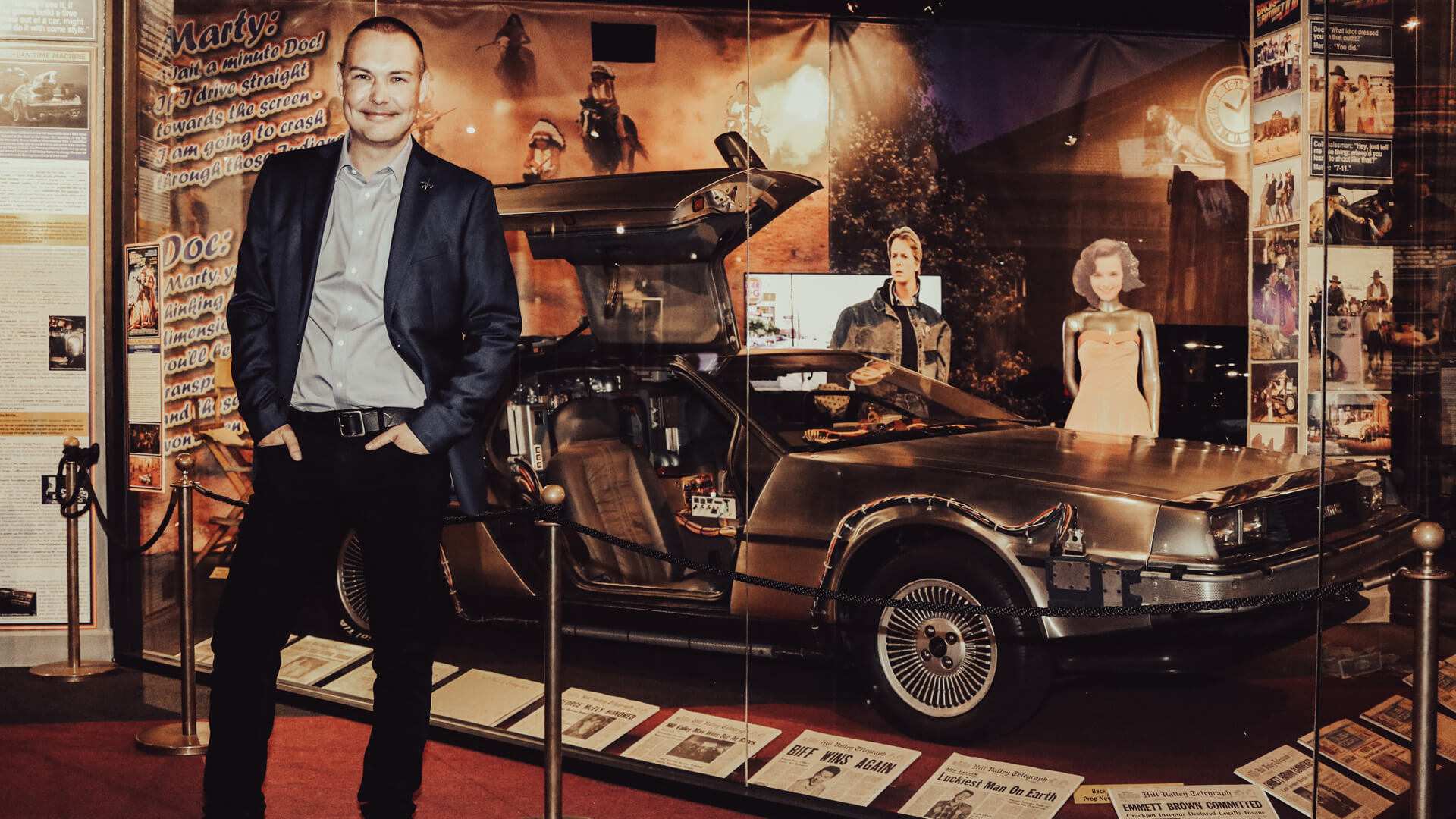A man in a suit stands beside a DeLorean among framed film memorabilia.