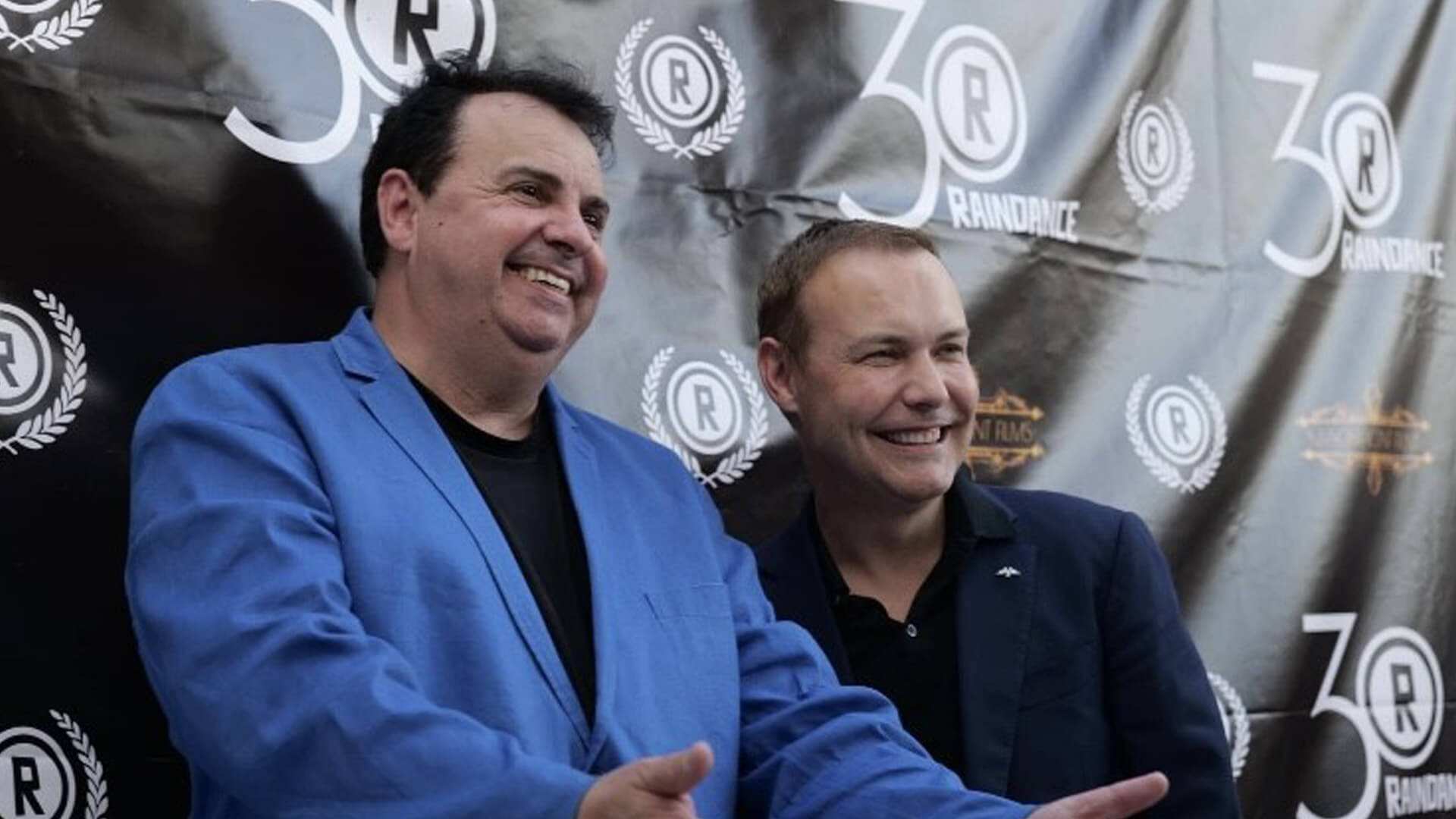Two men sit side by side, smiling, in front of a 30th anniversary backdrop.